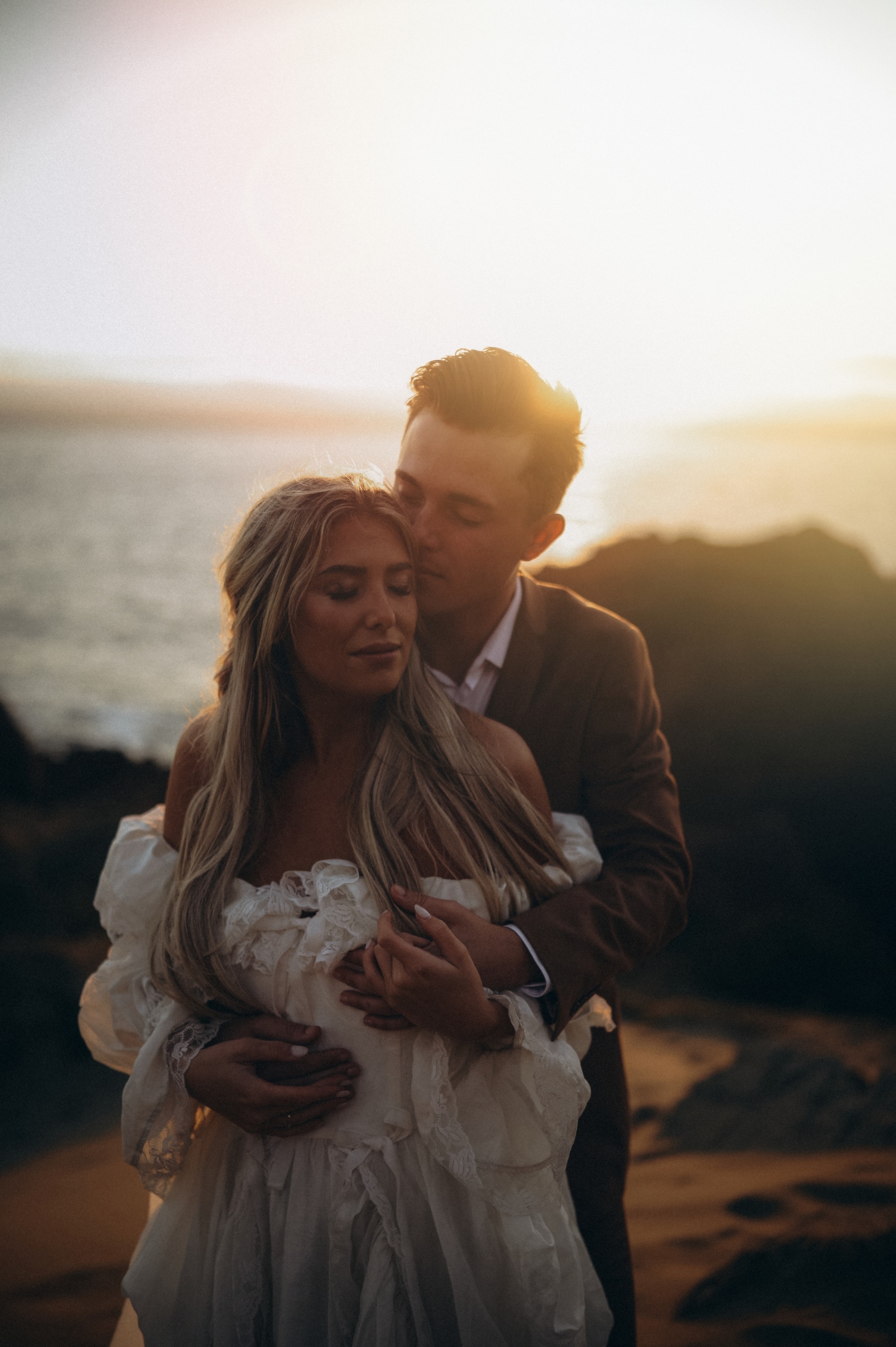 Blake holding Maria from behind on the bluff, her gown catching the sea wind