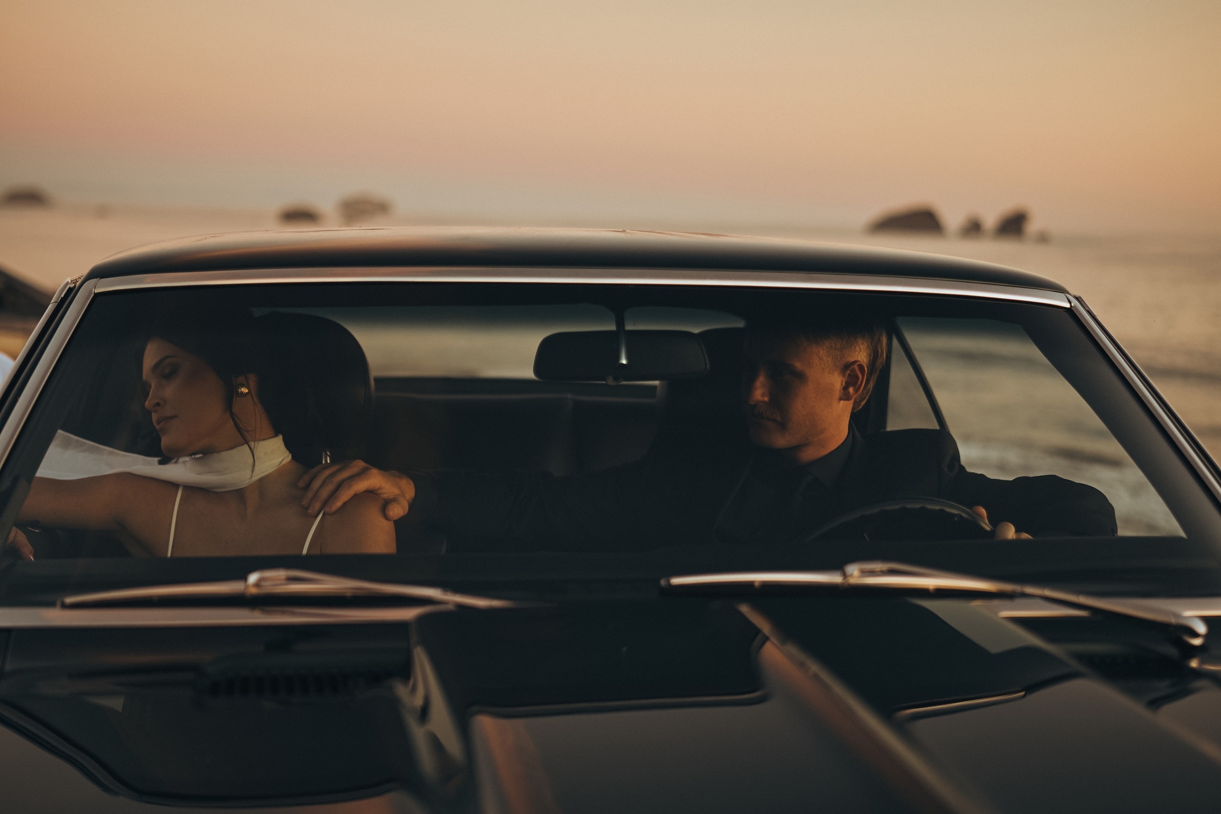 Bride and groom in a vintage car at coastal dusk, veil out the window