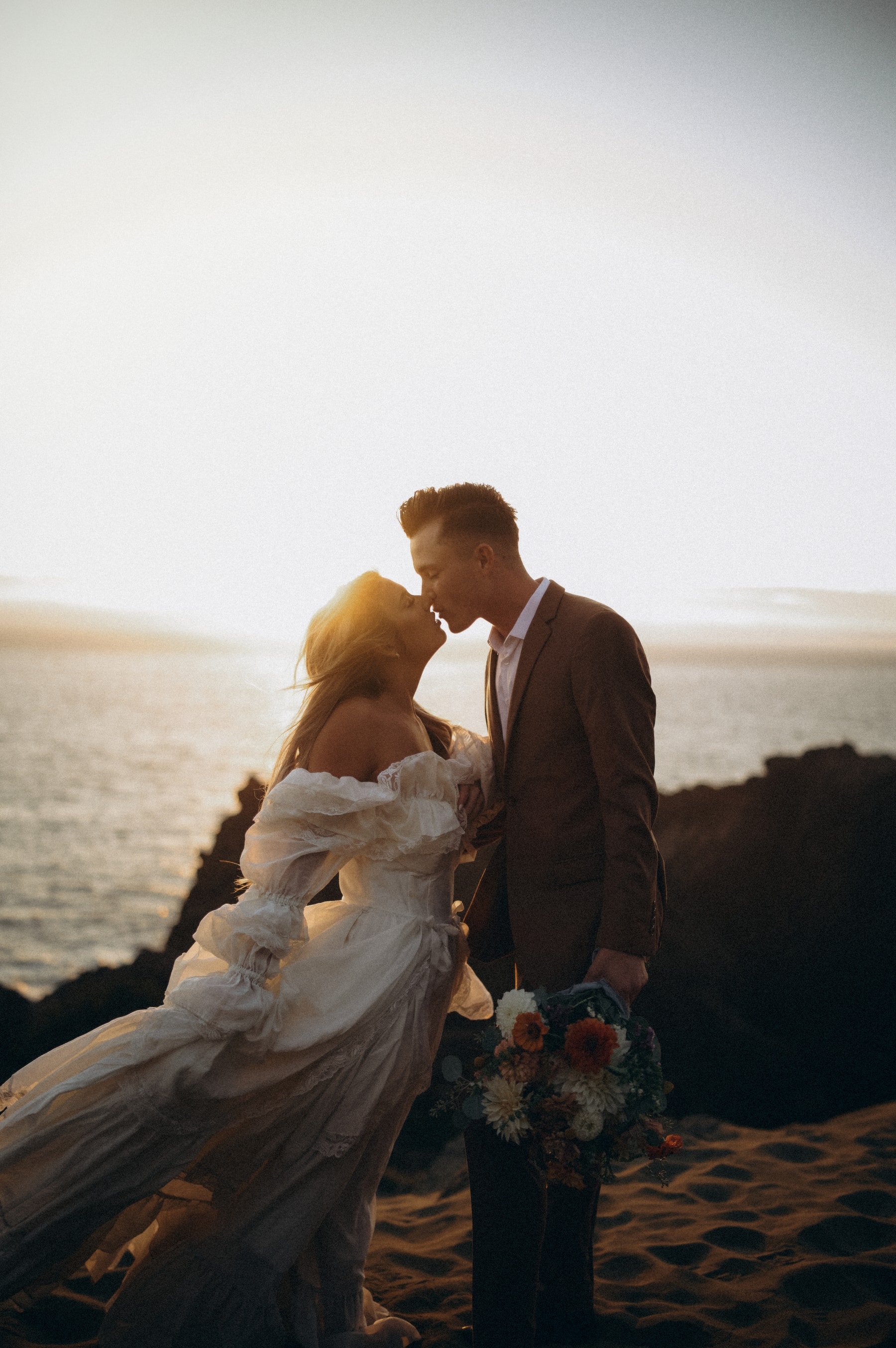 Bride and groom kissing at sunset on a coastal cliff, backlit by golden hour — fine art film photograph