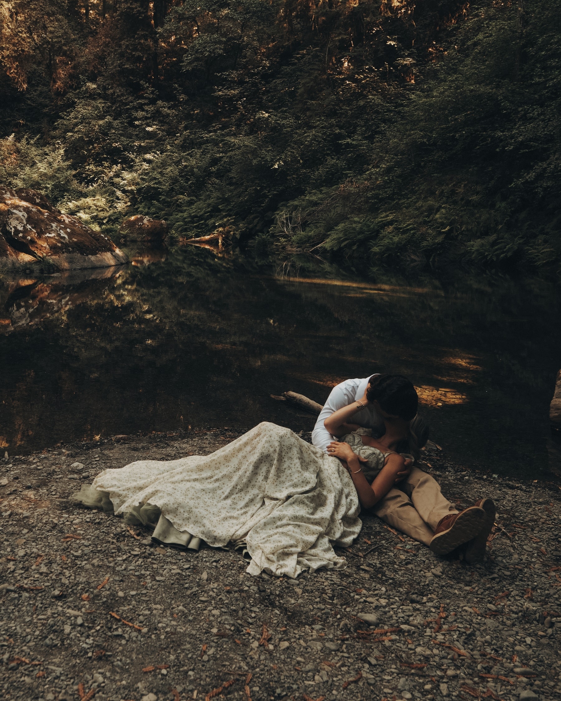 Blake and Maria lying beside a dark forest river beneath the redwoods, her gown spread across the gravel